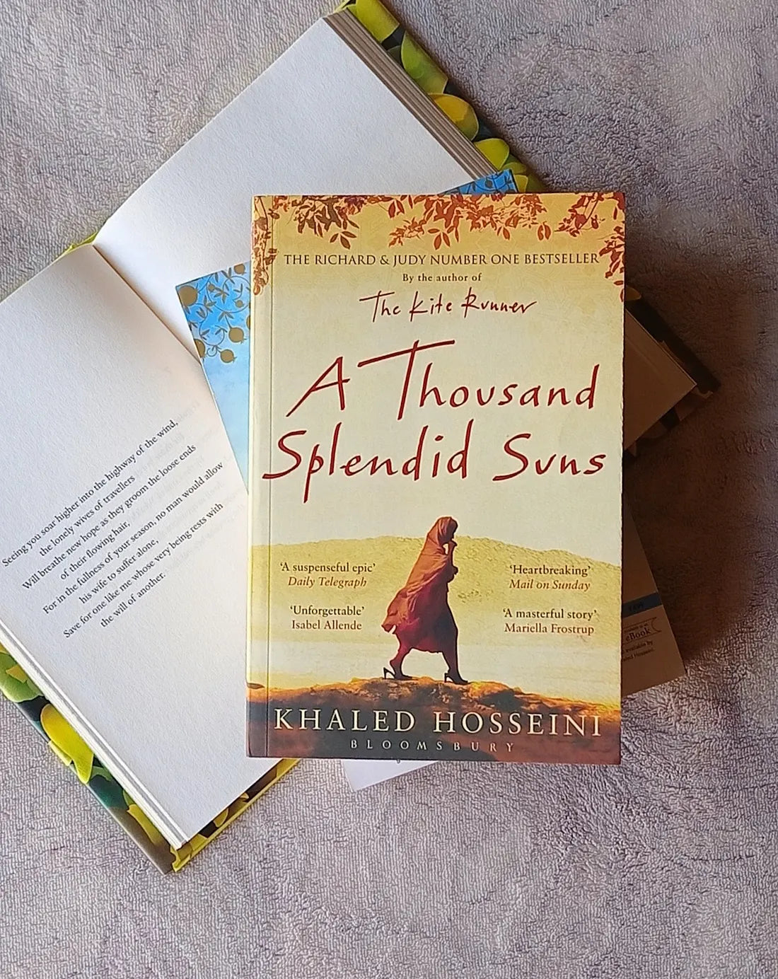 A book titled 'A Thousand Splendid Suns' by Khaled Hosseini lies open on a textured gray fabric surface, with its cover visible and another copy partially visible underneath.