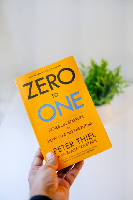 Book titled 'Zero to One' by Peter Thiel held by a hand with a plant in the background