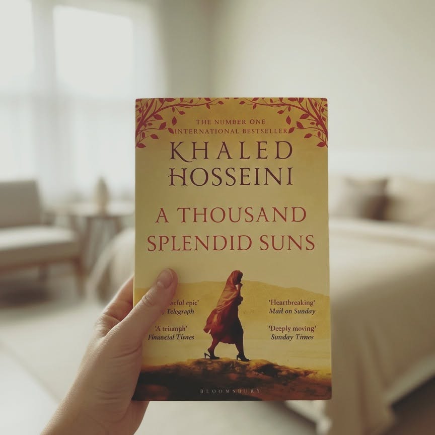 A hand holds a paperback copy of the book 'A Thousand Splendid Suns' by Khaled Hosseini. The book cover is a warm tan/beige color with dark brown and red text and illustrations, set against a softly blurred indoor background.