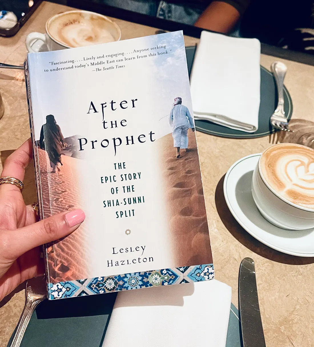 A hand holds a paperback copy of the book 'After the Prophet' by Lesley Hazleton, featuring a coffee table setting in the background with cappuccinos and cutlery.