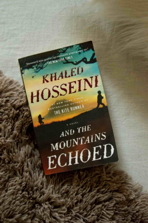 The book cover of 'And the Mountains Echoed' by Khaled Hosseini is shown resting on a brown shaggy rug over a light beige textured blanket.