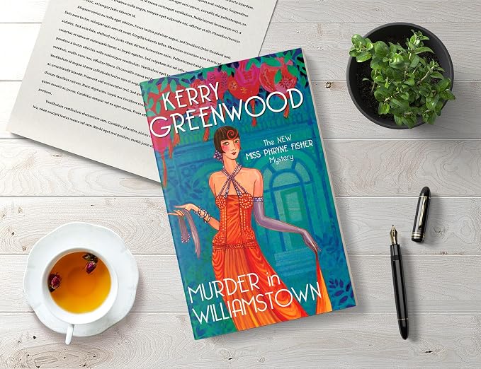 A book titled 'Murder in Williamstown' by Kerry Greenwood is displayed in a styled flat-lay on a white wooden surface, surrounded by a cup of tea, an open book, a small potted plant, and a pen.