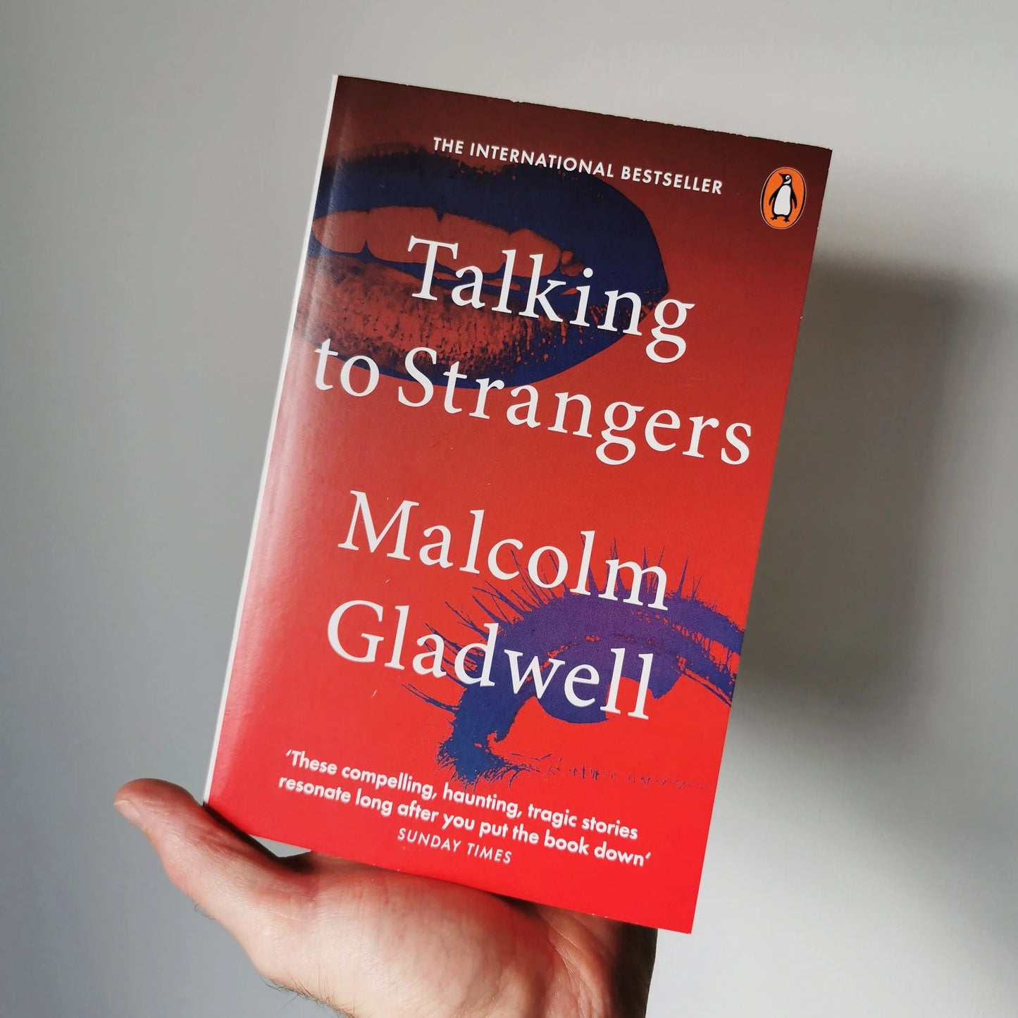 A hand holding a paperback copy of the book 'Talking to Strangers' by Malcolm Gladwell. The book cover is predominantly red with a dark blue and purple abstract design and white text.