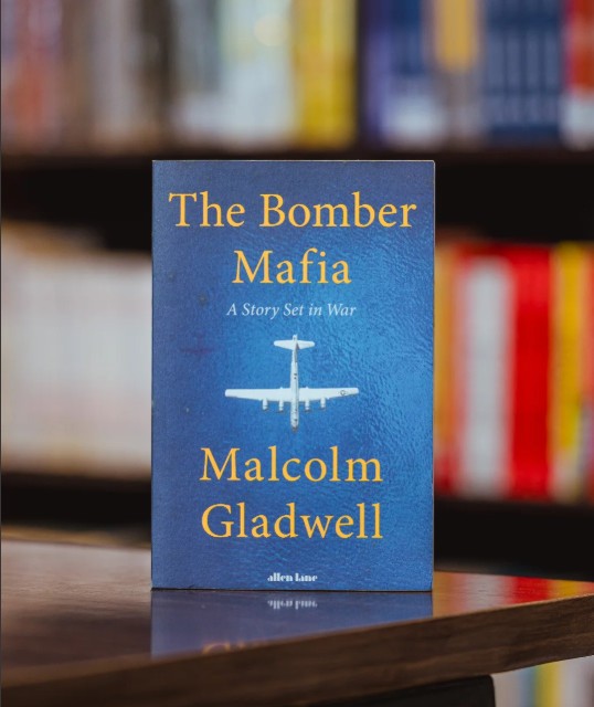 The book cover of 'The Bomber Mafia' by Malcolm Gladwell stands upright on a wooden shelf, with a blurred library or bookstore background of bookshelves and books.