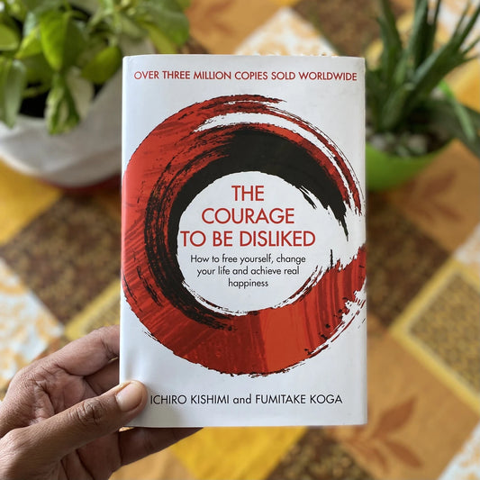 A hand holds a paperback book titled "The Courage to Be Disliked" by Ichiro Kishimi and Fumitake Koga. The book cover is white with a large red and black brushstroke design and red text.