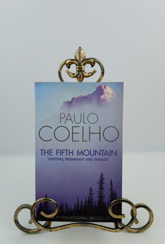 Book cover of Paulo Coelho's 'The Fifth Mountain' displayed upright in a decorative gold metal stand against a light gray background.