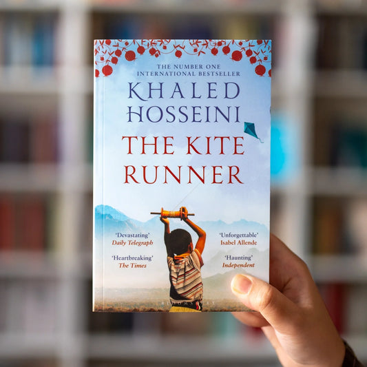 A hand holds a paperback copy of the book 'The Kite Runner' by Khaled Hosseini. The book cover features a light blue sky background with red decorative elements at the top, the title in large red letters, and an illustration of a boy flying a kite near the bottom.