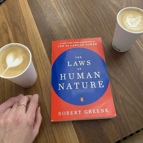 A copy of the book 'The Laws of Human Nature' by Robert Greene lies on a wooden table, flanked by two cups of coffee with latte art. A hand is visible in the bottom-left corner.