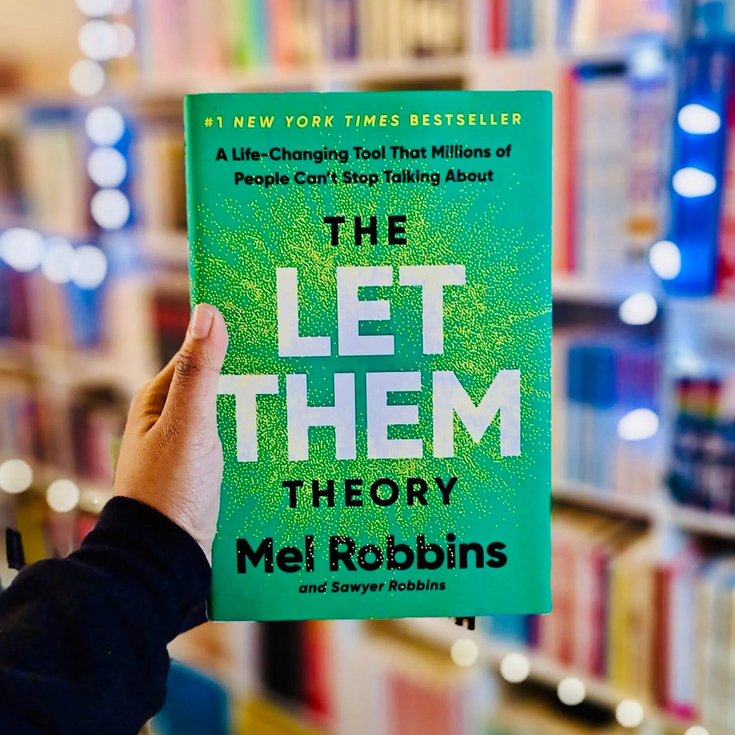 A hand holds a bright green copy of the book 'The Let Them Theory' by Mel Robbins in front of a blurred bookstore background with bookshelves and warm string lights.