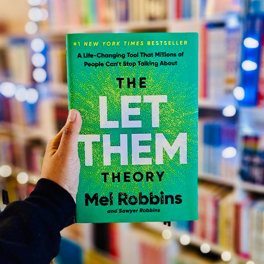 A hand holds a bright green copy of the book 'The Let Them Theory' by Mel Robbins in front of a blurred bookstore background with bookshelves and warm string lights.