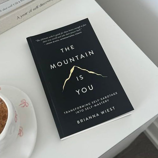 Book titled 'The Mountain is You' by Brianna West on a table with a cup of coffee and books in the background.