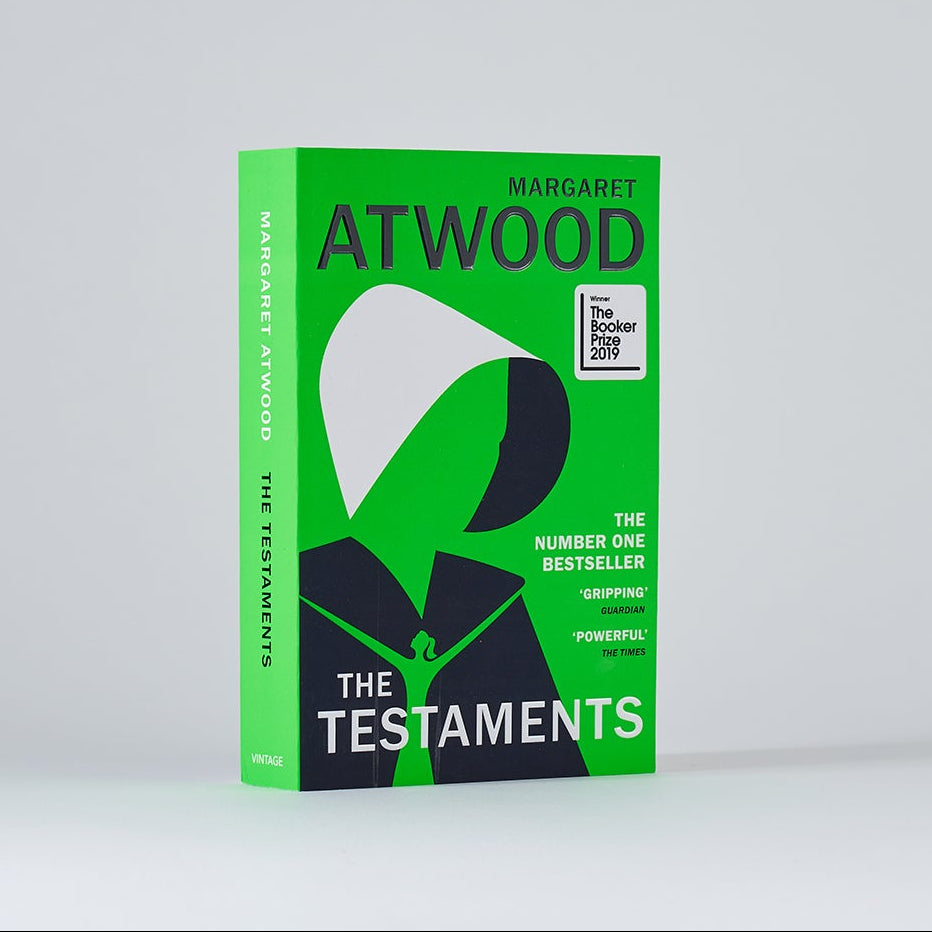 The book 'The Testaments' by Atwood, featuring a bright green cover with a stylized black and white hooded figure, photographed on a light gray background.