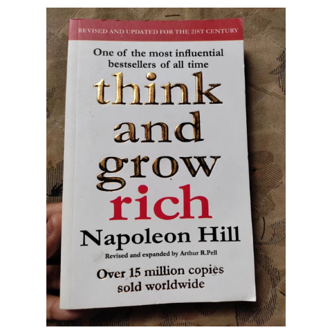 A paperback copy of the book 'Think and Grow Rich' by Napoleon Hill, held in a hand against a textured brown background. The cover is white with metallic gold and red lettering.